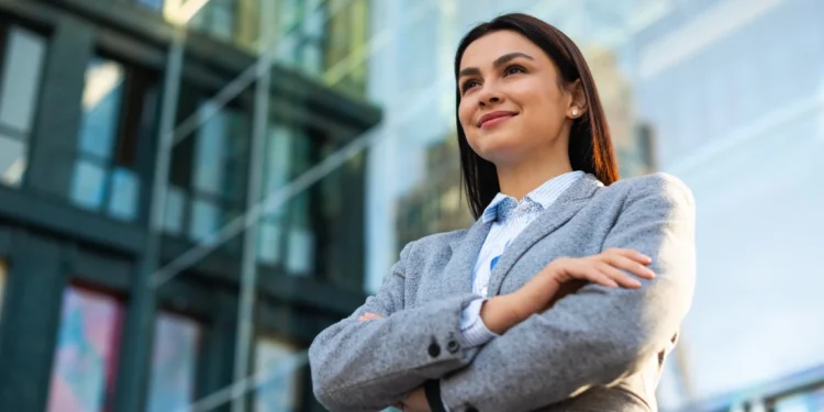 Mulher confiante em traje de negócios, posando com os braços cruzados, olhando com um leve sorriso em um cenário urbano moderno.