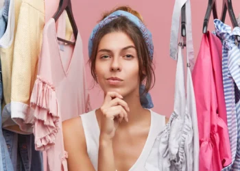 Mulher pensativa, usando um lenço na cabeça, rodeada por roupas coloridas em um guarda-roupa, refletindo sobre o que escolher para criar novos looks.