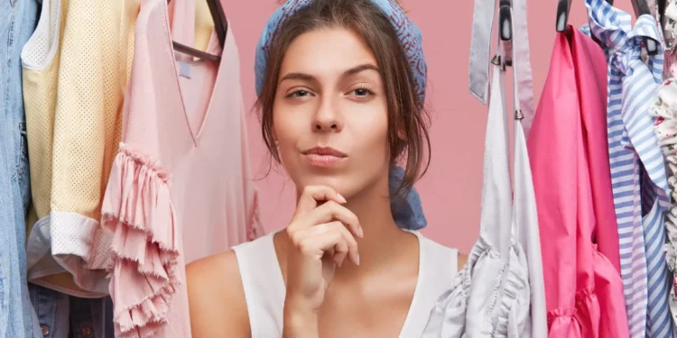 Mulher pensativa, usando um lenço na cabeça, rodeada por roupas coloridas em um guarda-roupa, refletindo sobre o que escolher para criar novos looks.