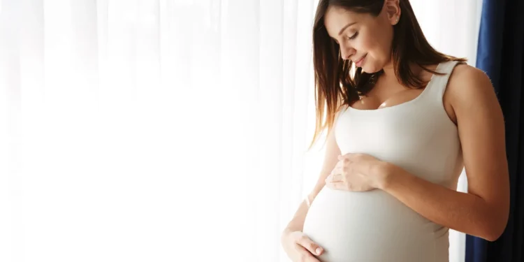 Mulher grávida tocando sua barriga com um sorriso suave, refletindo felicidade e tranquilidade na sua primeira gestação.