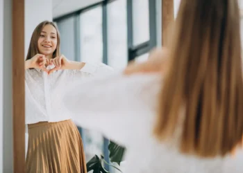 Mulher jovem sorrindo para o espelho, fazendo um coração com as mãos enquanto veste uma camisa branca e saia dourada, refletindo sobre a importância do autocuidado.