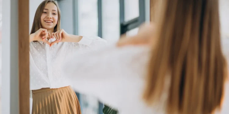 Mulher jovem sorrindo para o espelho, fazendo um coração com as mãos enquanto veste uma camisa branca e saia dourada, refletindo sobre a importância do autocuidado.