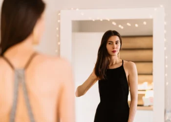 Como se sentir linda em qualquer momento, mesmo nos dias difíceis. Mulher refletindo no espelho com um vestido preto, simbolizando autoestima e confiança.