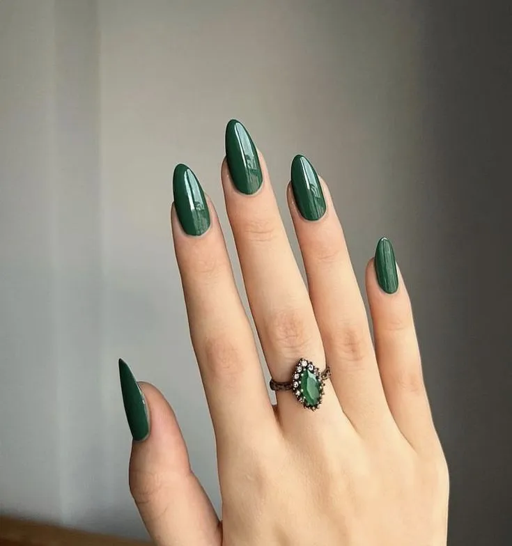 Imagem de unha pintada com verde musgo, com anel de pedra verde, transmitindo sofisticação e elegância.
