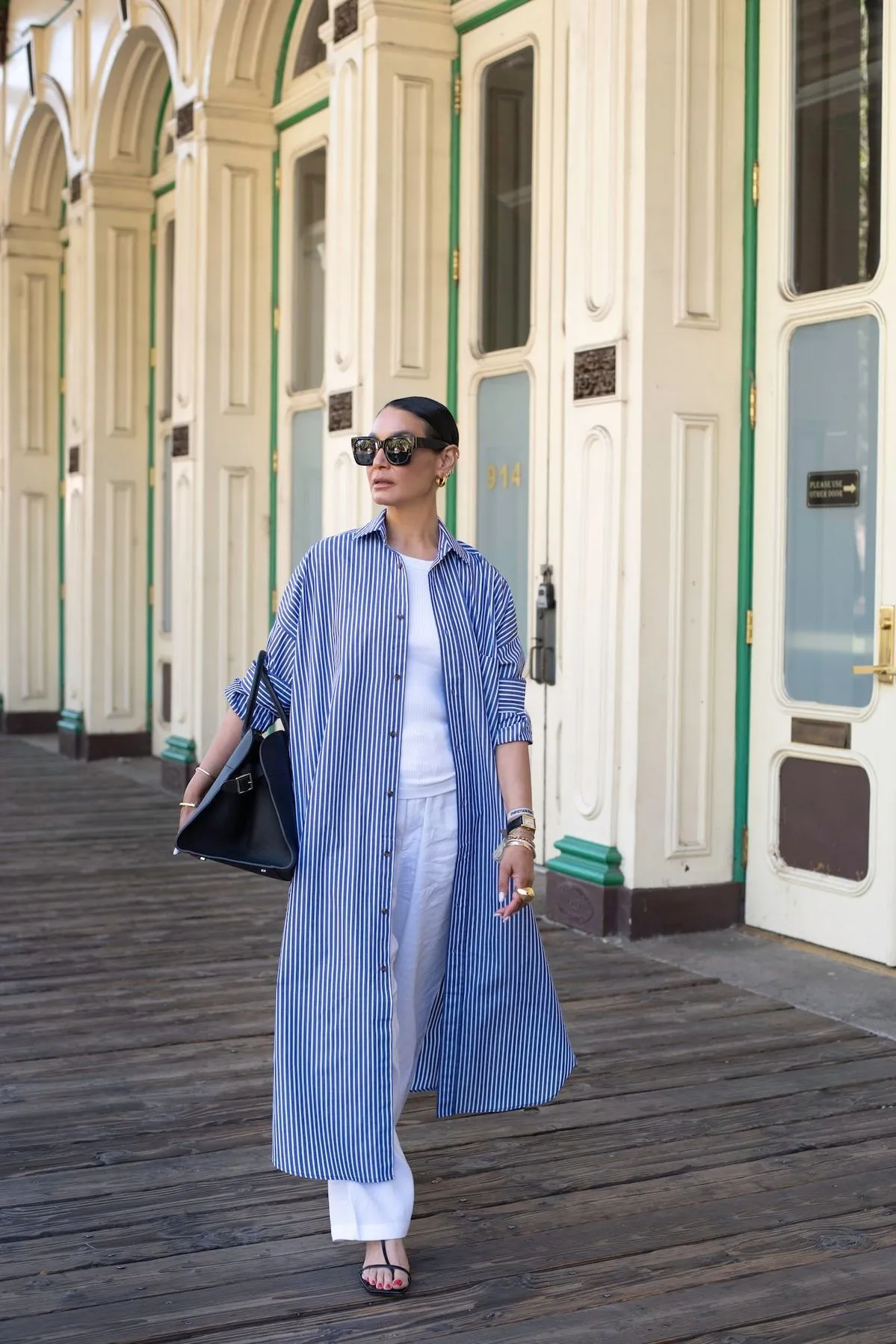 Mulher usando um vestido chemise listrado azul e branco, com óculos de sol e bolsa preta, em um ambiente ao ar livre.