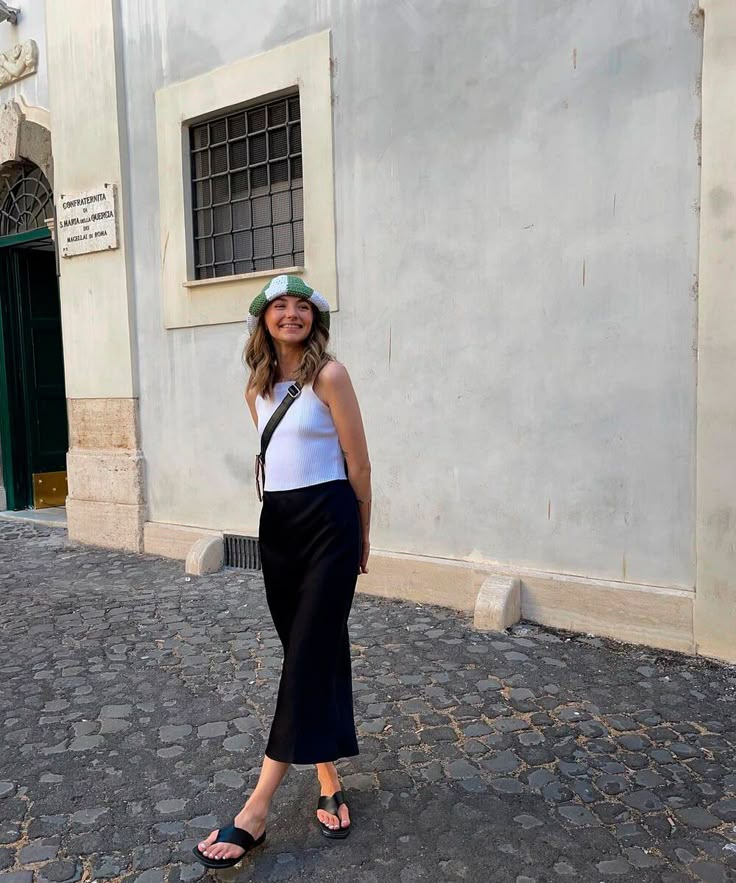 A imagem de uma mulher com um look casual de saia preta e regata branca, usando chinelos pretos e um chapéu colorido.