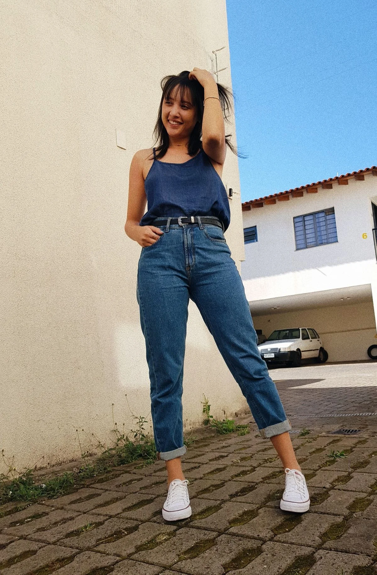 Mulher usando all star branco, mom jeans e blusa azul, sorrindo ao lado de uma parede e com céu azul ao fundo.