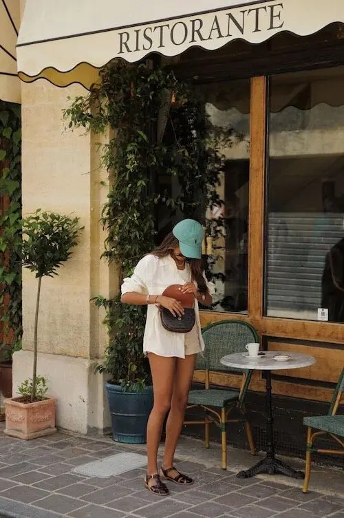 Mulher com boné e roupas leves, parada em frente a um restaurante, ajustando sua bolsa, em uma cena de verão.