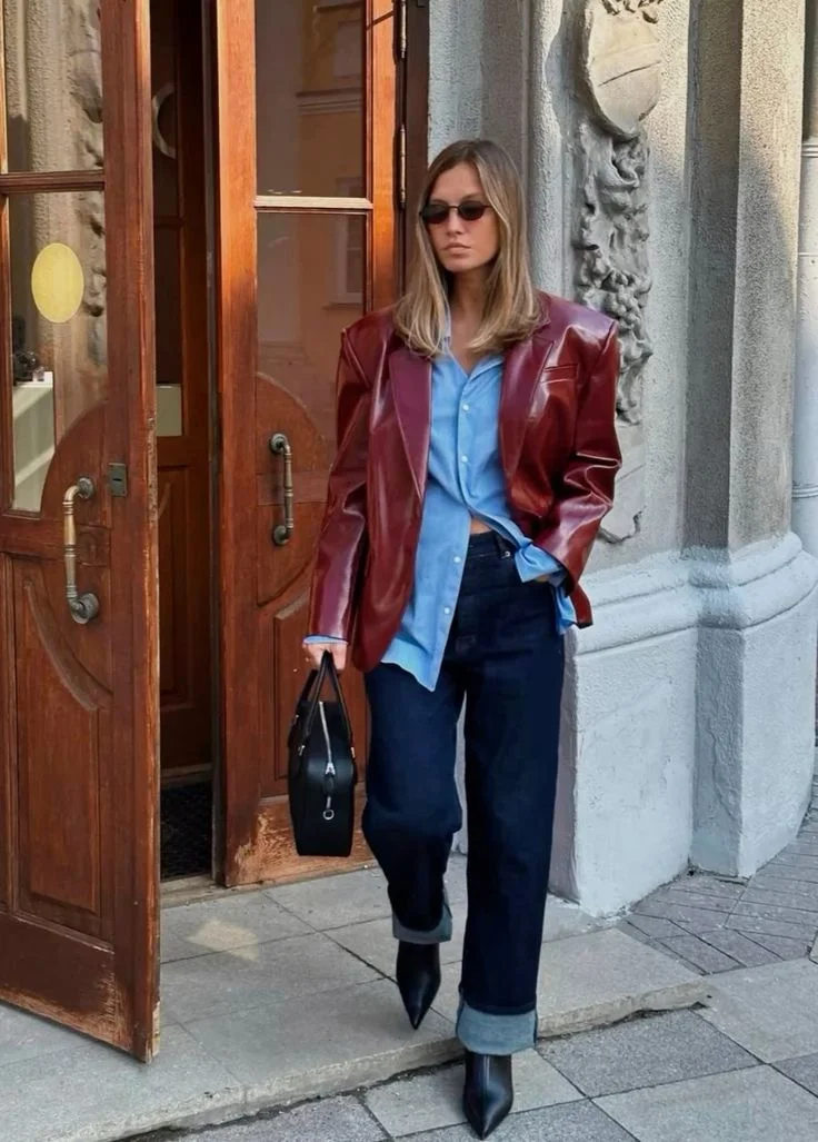 Pessoa usando camisa social azul clara com jaqueta de couro marrom e calças escuras, andando na rua com bolsa preta e sapatos de salto.