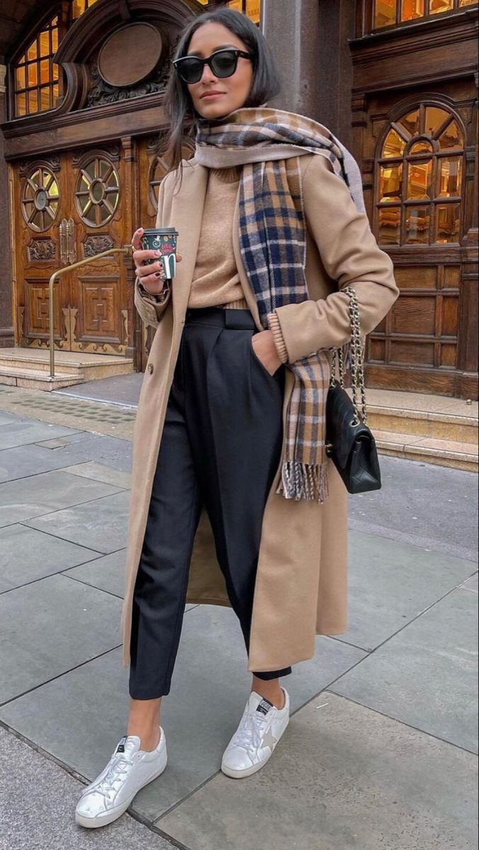 Mulher vestindo um look de inverno elegante, com casaco longo, calça de alfaiataria preta, suéter e cachecol xadrez.