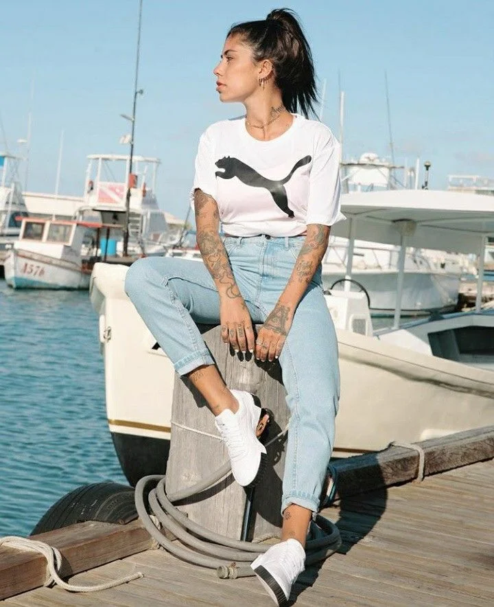 Imagem de uma jovem usando um Puma Cali branco, com uma camiseta branca e calças jeans claras, sentada em um cais próximo a barcos.