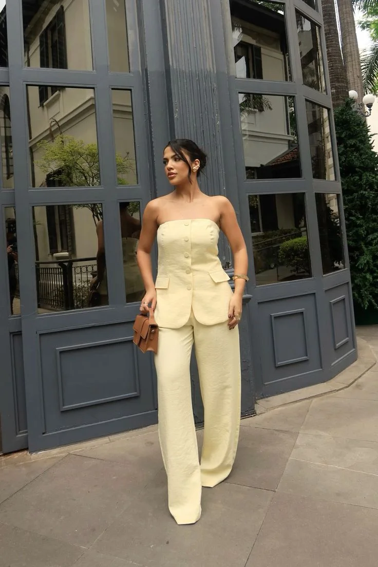 Mulher com conjunto amarelo manteiga, top sem alças e calça de corte reto, posando ao lado de uma janela.