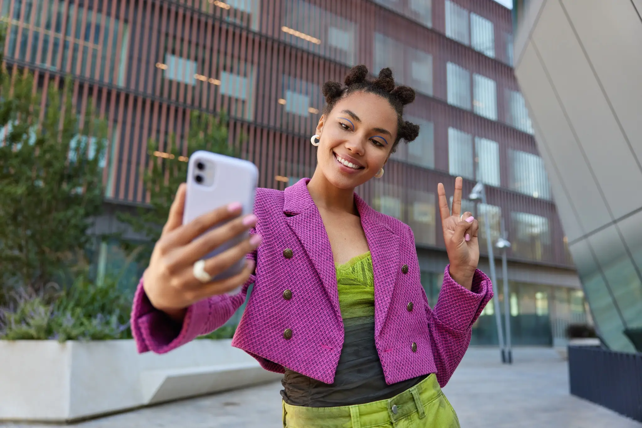Mulher sorrindo e fazendo sinal de paz enquanto tira uma selfie ao ar livre, com roupas coloridas e estilo moderno.