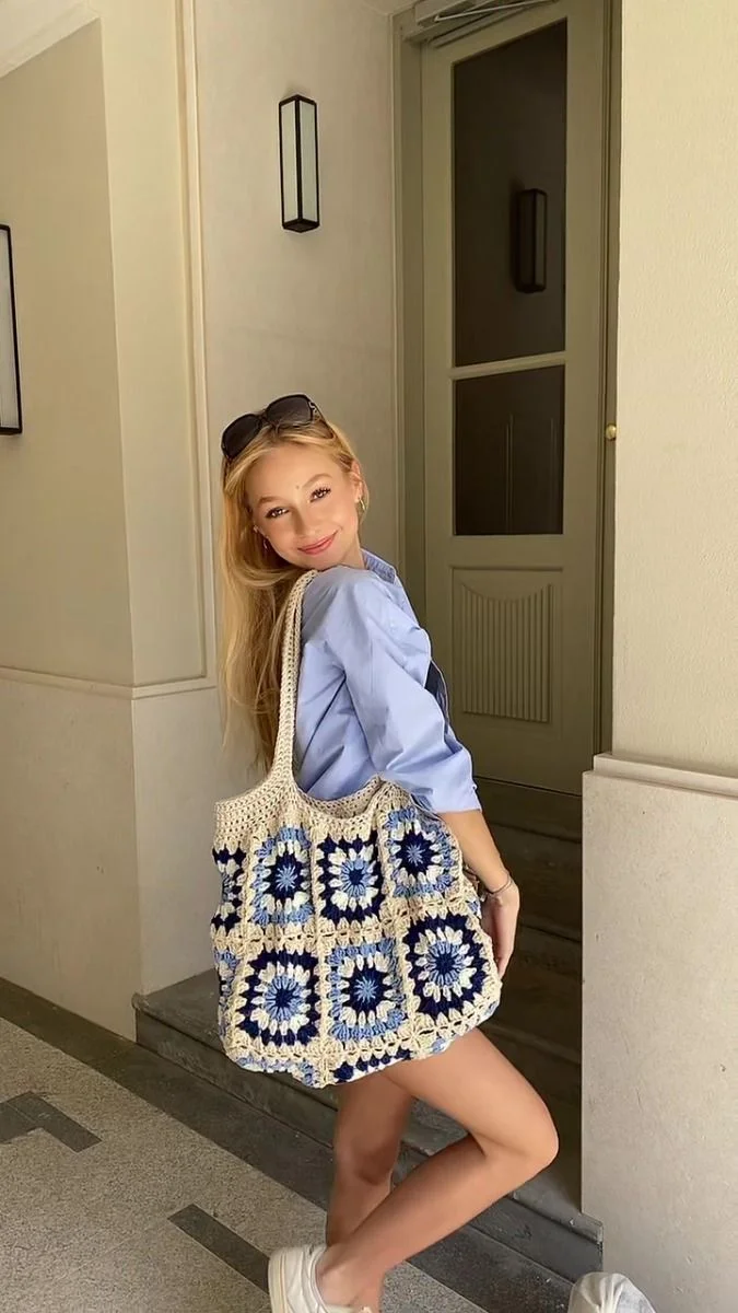 Mulher usando camisa azul e short, segurando bolsa de crochê com detalhes em azul e branco.