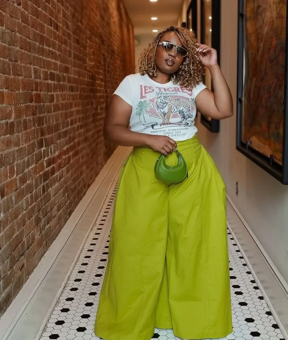 Mulher usando calça balloon verde com camiseta estampada e acessórios coloridos, em um ambiente moderno e descontraído.