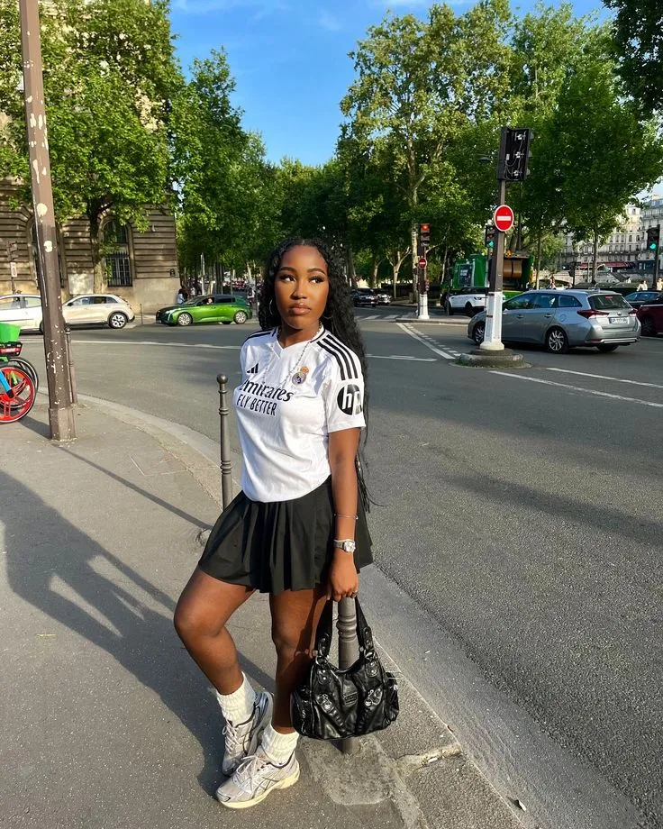 Mulher usando camisa de time e saia midi plissada, com tênis e bolsa, em um ambiente urbano.