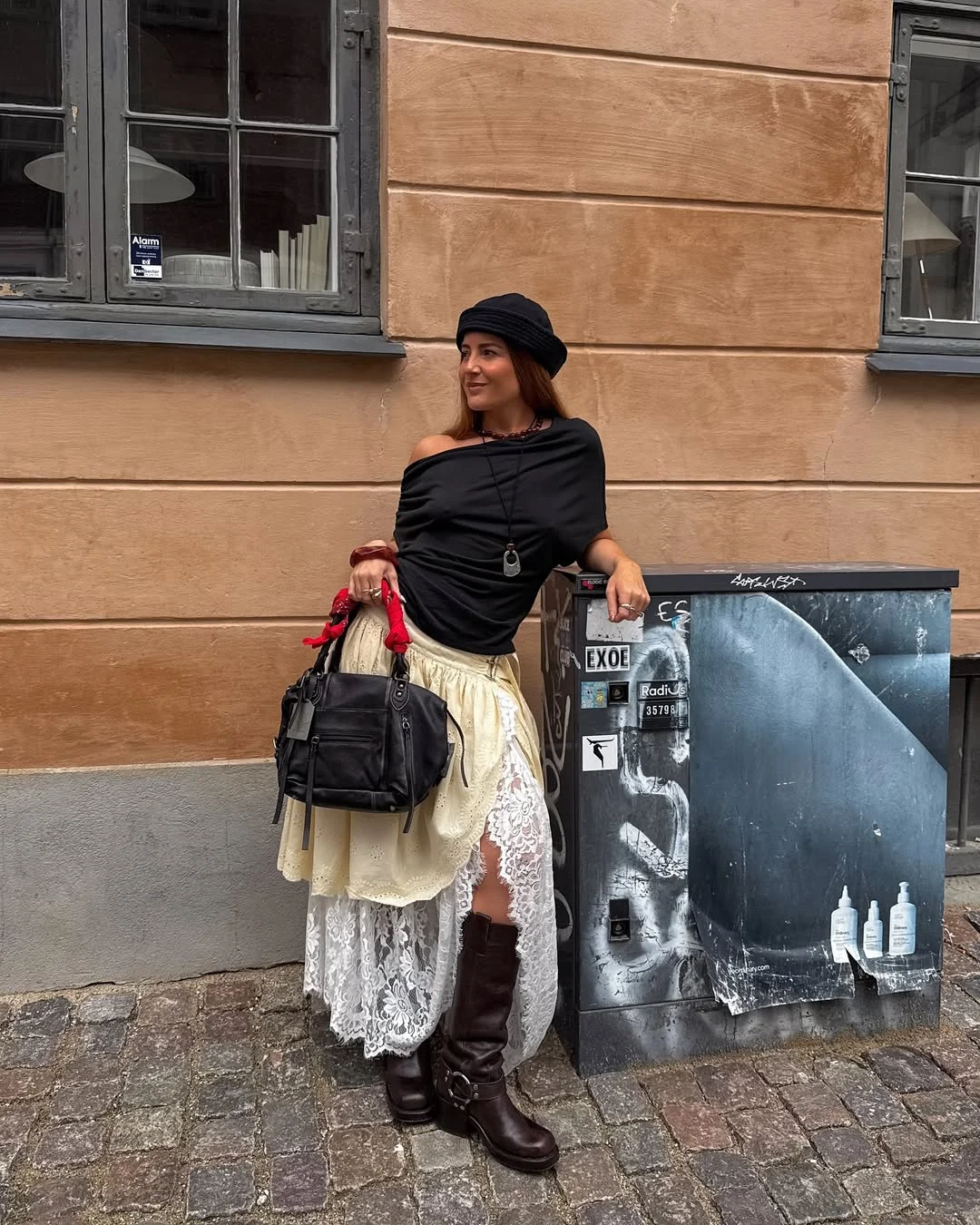 Mulher estilosa posando na rua, com um chapéu preto e uma blusa de manga curta, usando botas e uma saia com renda, em um cenário urbano.