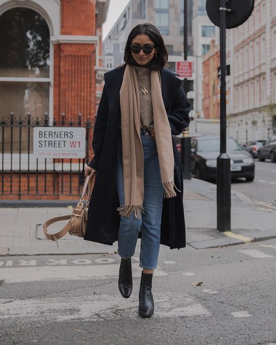 Mulher atravessando a rua com sobretudo azul marinho, jeans, botas pretas e um longo cachecol bege.