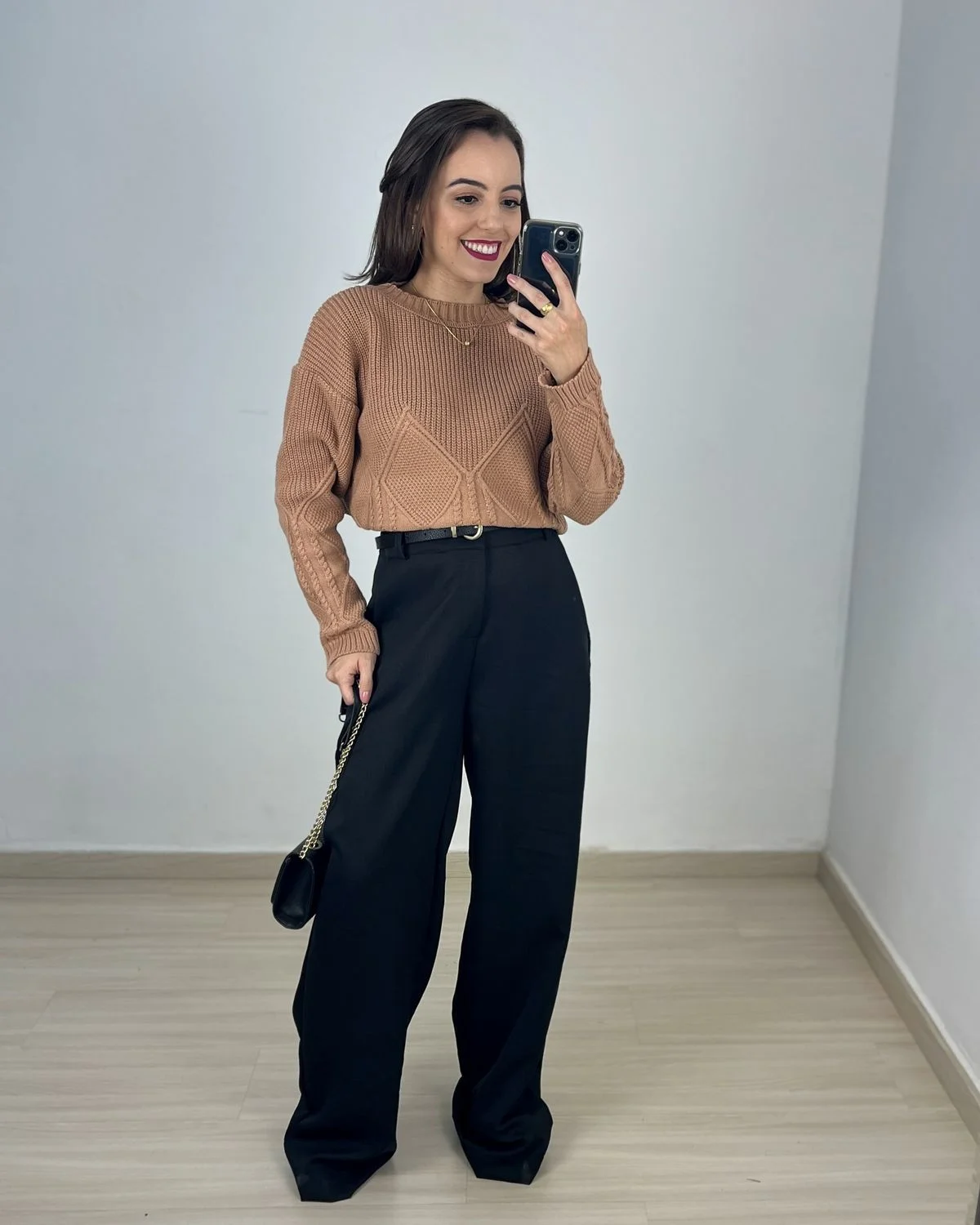 Mulher sorrindo e usando um visual elegante com suéter de tricô bege e calça preta de corte reto, em um ambiente neutro.