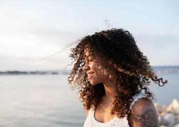 Mulher com cabelos cacheados sorrindo à beira-mar, aproveitando a luz suave do entardecer