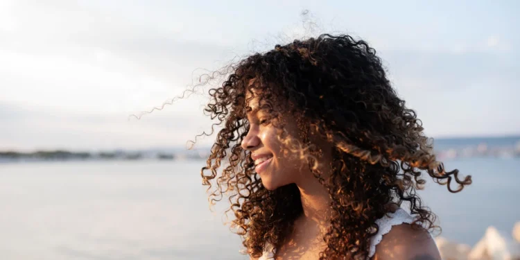 Cabelos cacheados: 5 truques para definir e hidratar os fios 1 Mulher com cabelos cacheados sorrindo à beira-mar, aproveitando a luz suave do entardecer
