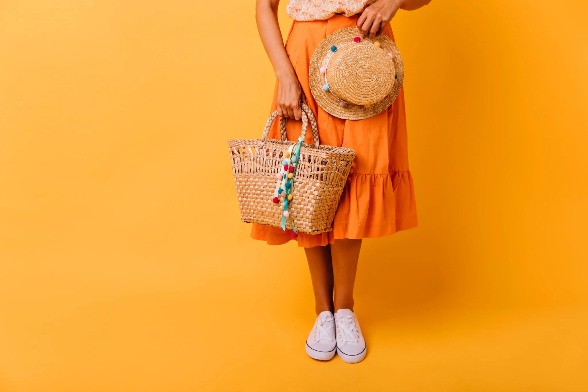 Mulher com saia laranja, tênis branco e acessórios de palha, incluindo bolsa e chapéu colorido