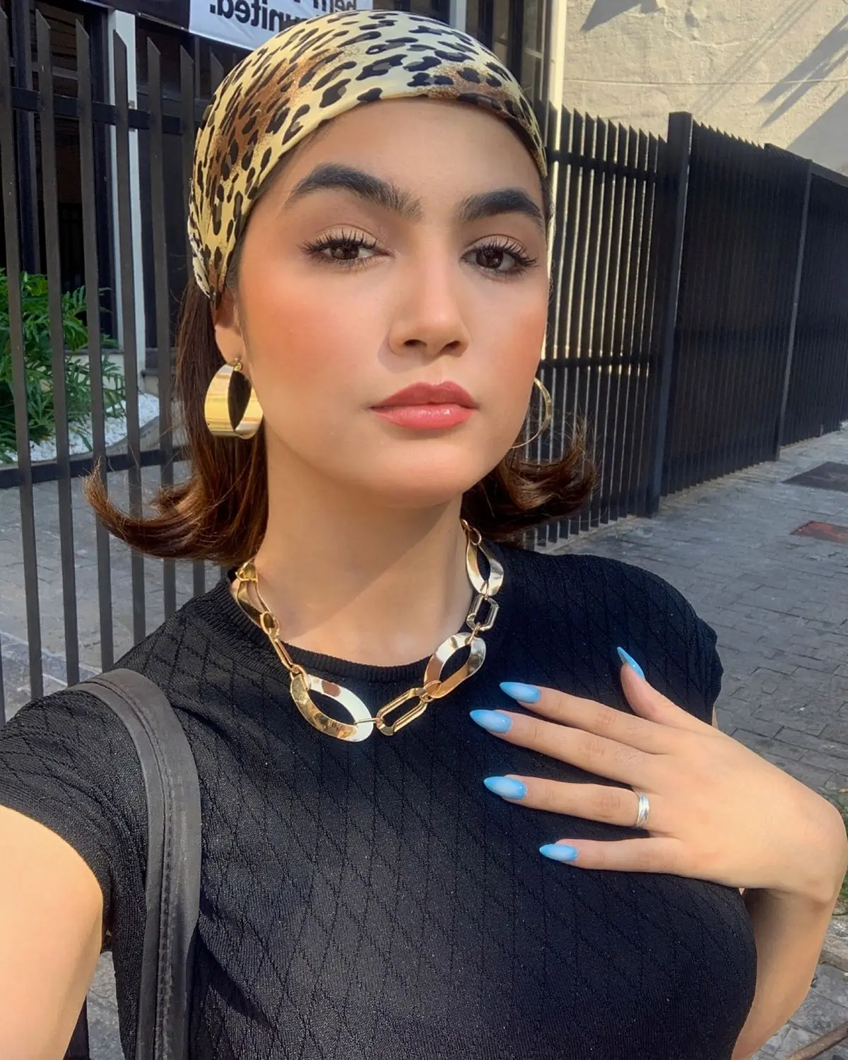 Mulher com lenço de estampa de onça no cabelo, usando blusa preta e acessórios dourados, posando para foto ao ar livre.