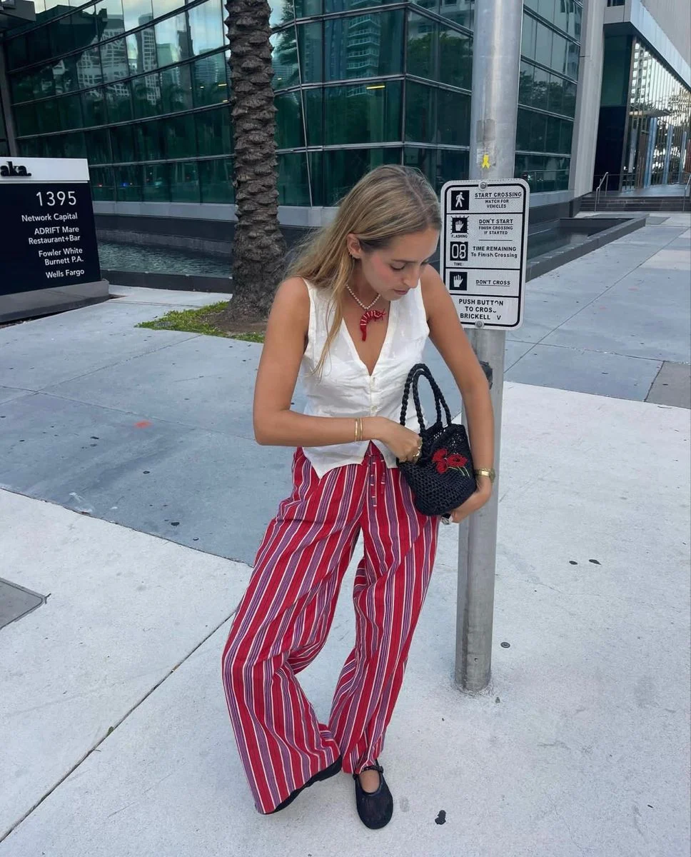 Mulher usando blusa branca e calça listrada vermelha e branca com bolso e bolsa preta, caminhando na rua.