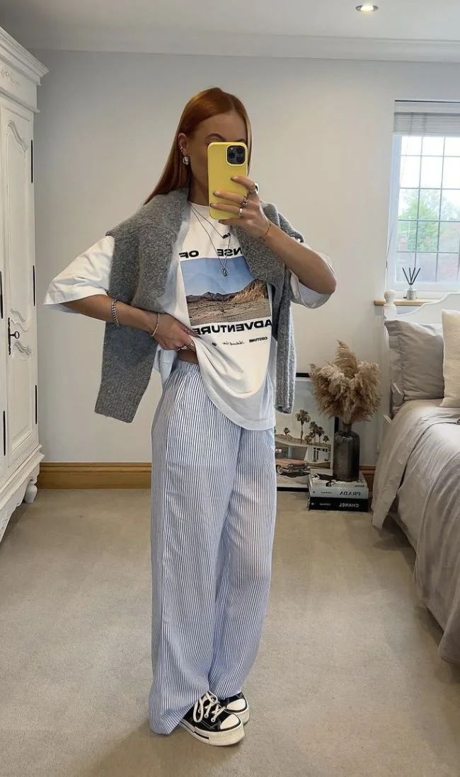 Pessoa posando com um look casual, composto por uma camiseta branca com estampa, colete cinza, calça listrada e tênis Converse.