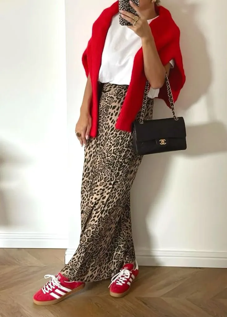 Look básico com estampa de leopardo, composto por uma blusa branca, saia longa com estampa animal print e tênis vermelho Adidas.