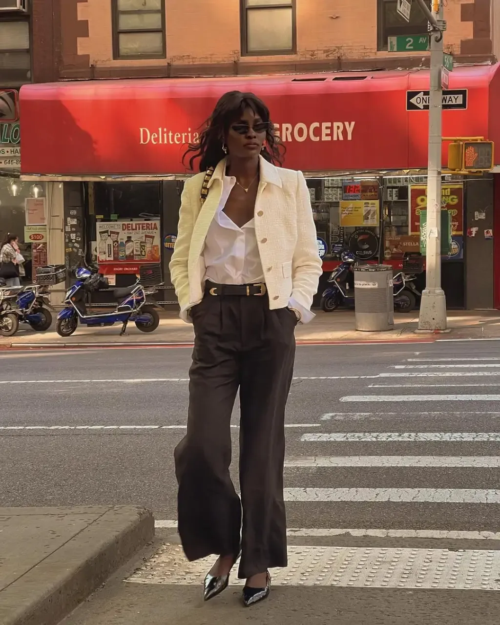 Imagem de mulher estilosa com camisa social branca e jaqueta amarela manteiga, calça preta e salto alto, em uma rua urbana.