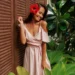 8 tendências da Primavera Verão para 2025 que você vai querer usar 13 Mulher sorridente usando vestido rosa com detalhes delicados, com flor de hibisco no cabelo, em ambiente tropical.