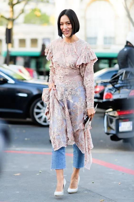 Mulher com vestido de renda rosa claro e jeans, posando para foto, usando salto branco, no estilo street style.