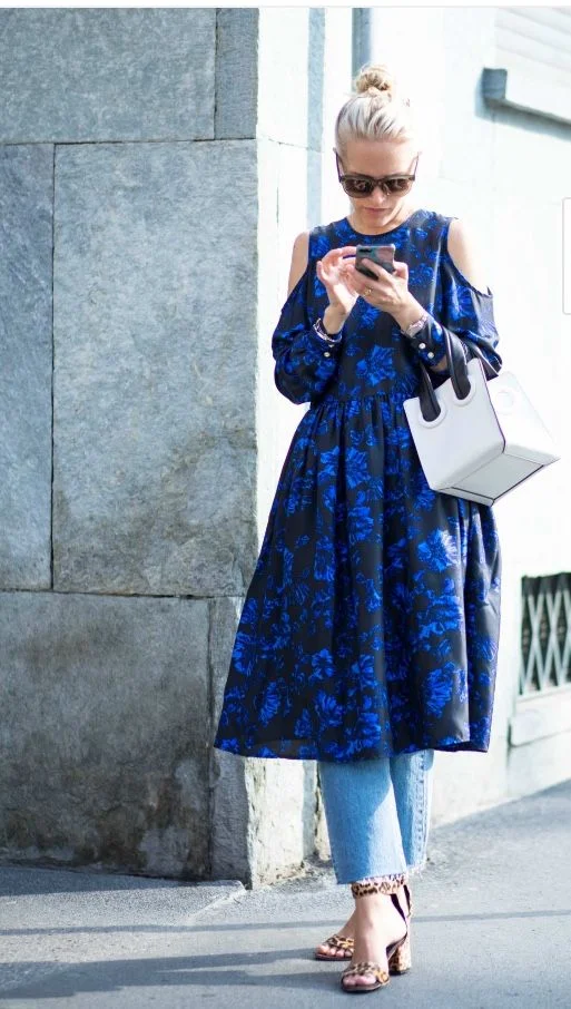 Mulher com vestido azul florido e calça jeans, usando óculos de sol e sandálias de salto alto com estampa de onça, checando o celular.