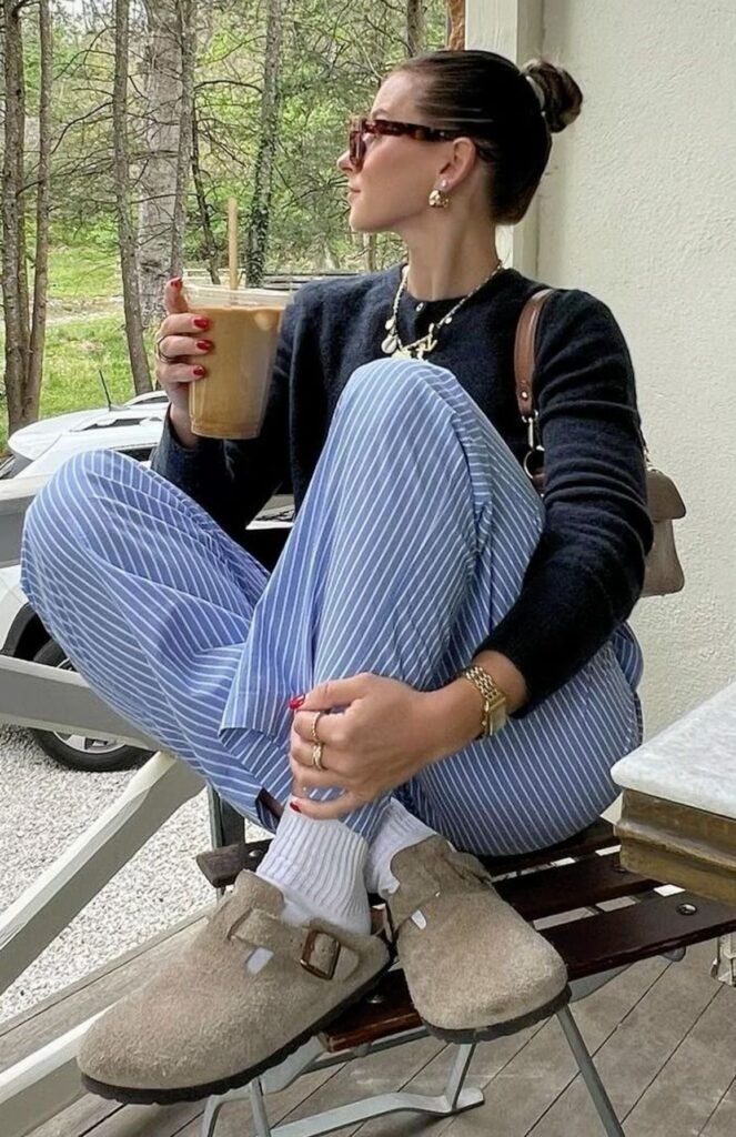 Mulher sentada em uma varanda, usando um par de tamancos clogs de camurça bege, calças listradas azuis e um suéter preto, exemplificando o estilo boho confortável.