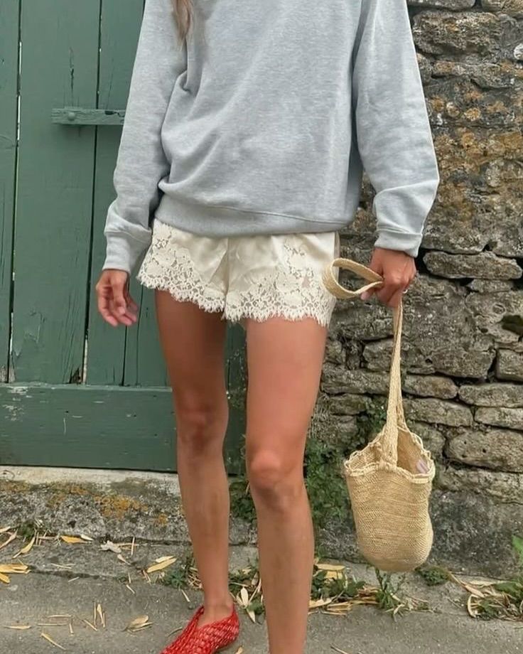 Mulher vestindo um short de cetim off-white com barra de renda, suéter cinza e sapatilhas vermelhas, segurando uma bolsa de palha.