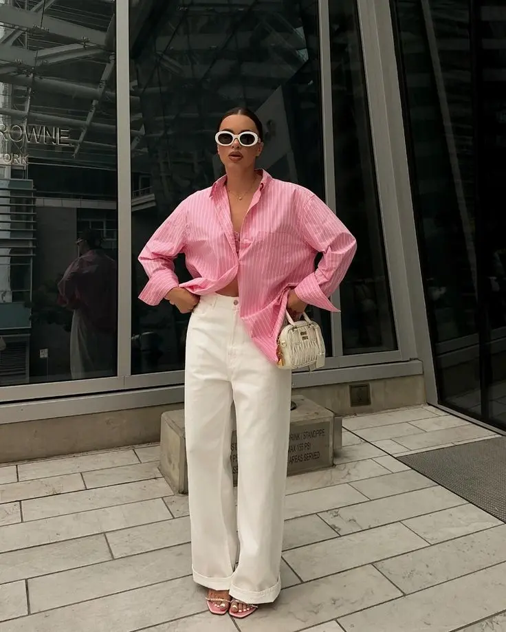 Look com camisa de listras rosa e branca, com um botão fechado, calça branca de cintura alta e óculos de sol, em ambiente urbano.