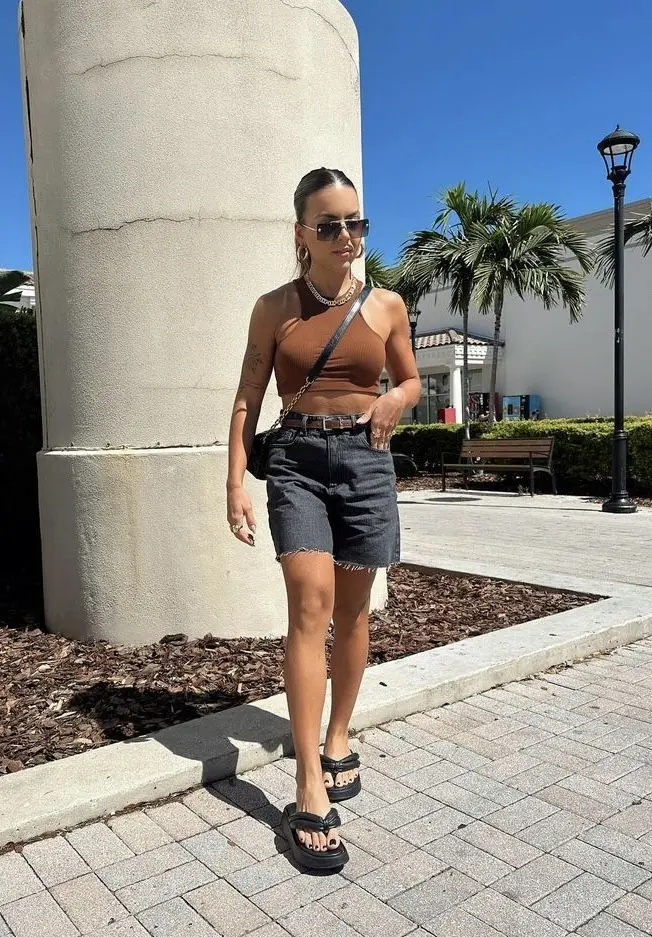 Mulher usando um top marrom, shorts jeans e chinelos de dedo, caminhando em uma área externa.