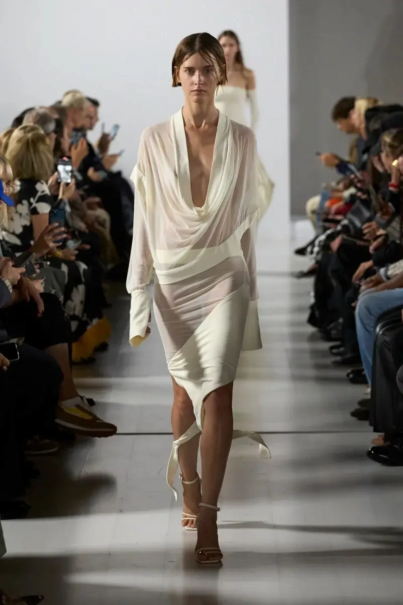 Modelo desfilando com vestido fluido branco com drapeados, apresentando fluidez estratégica na coleção SS26.