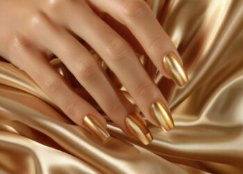 Mão feminina com unhas pintadas de esmalte metalizado dourado sobre tecido acetinado dourado