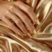 Brilho e estilo: 10 esmaltes metalizados para arrasar no visual 14 Mão feminina com unhas pintadas de esmalte metalizado dourado sobre tecido acetinado dourado