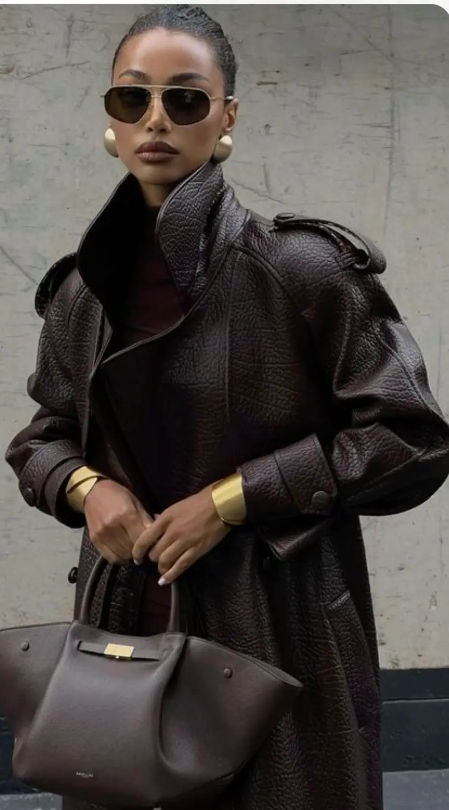 Mulher elegante usando sobretudo de couro marrom com gola alta, bolsa estruturada e óculos escuros.