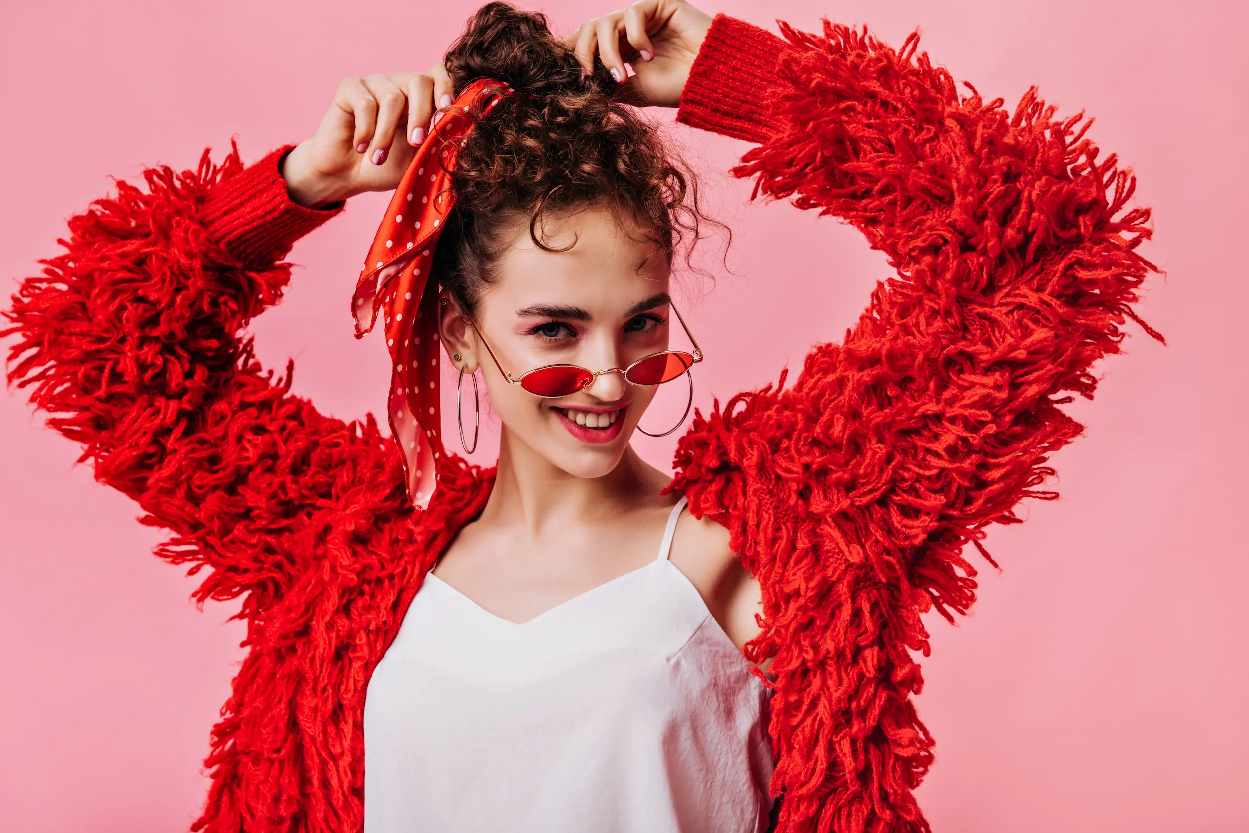 Mulher sorrindo usando um suéter vermelho, óculos de sol e lenço vermelho, em um fundo rosa. As franjas da peça adicionam movimento e fluidez ao visual.
