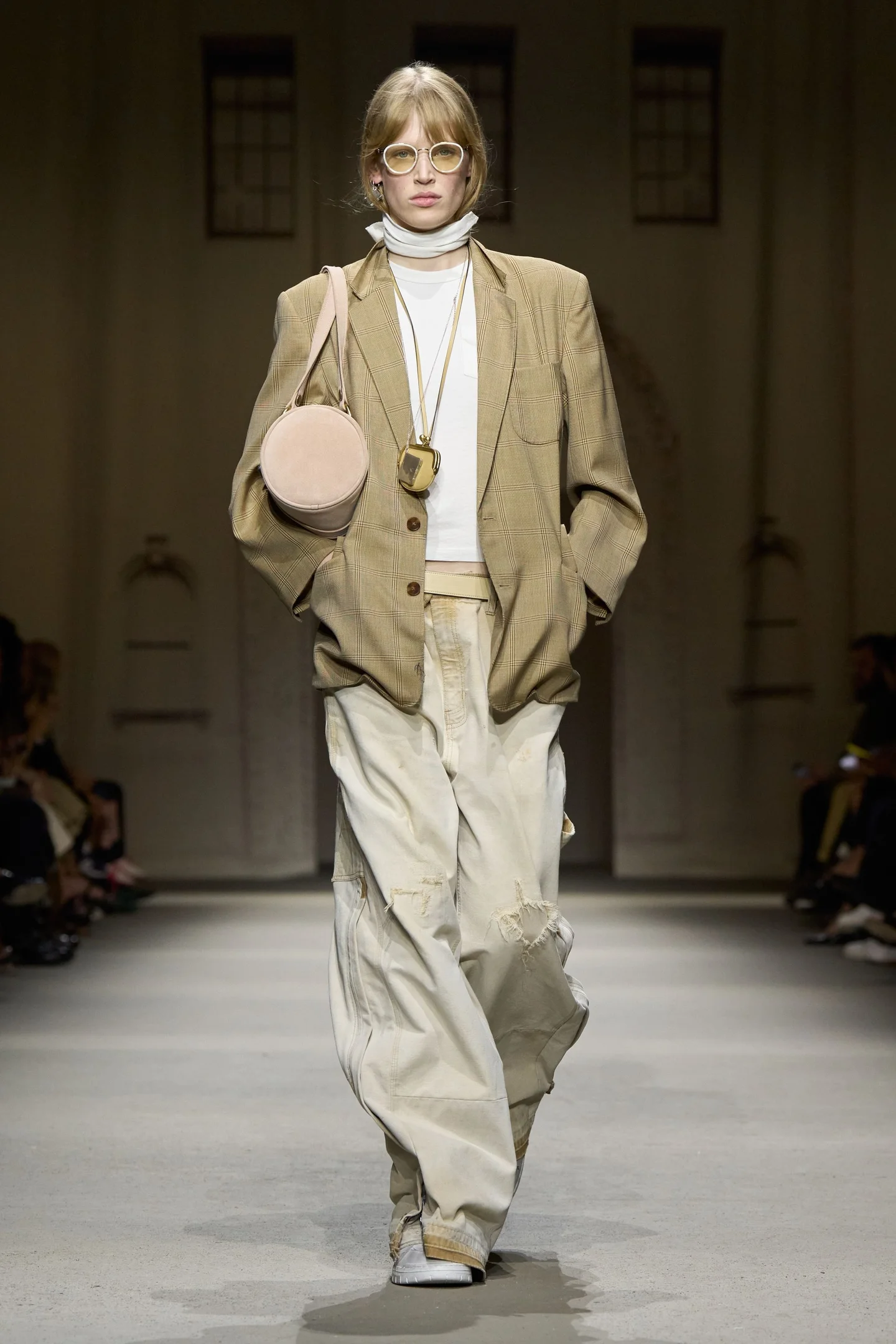 Modelo desfilando com visual prático e moderno, com blazer xadrez, calças largas e acessórios multifuncionais como uma bolsa que também funciona como um colar, representando a tendência de praticidade da New York Fashion Week SS26.