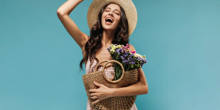 Mulher sorrindo com flores na cesta, vestindo chapéu, simbolizando a chegada da primavera.