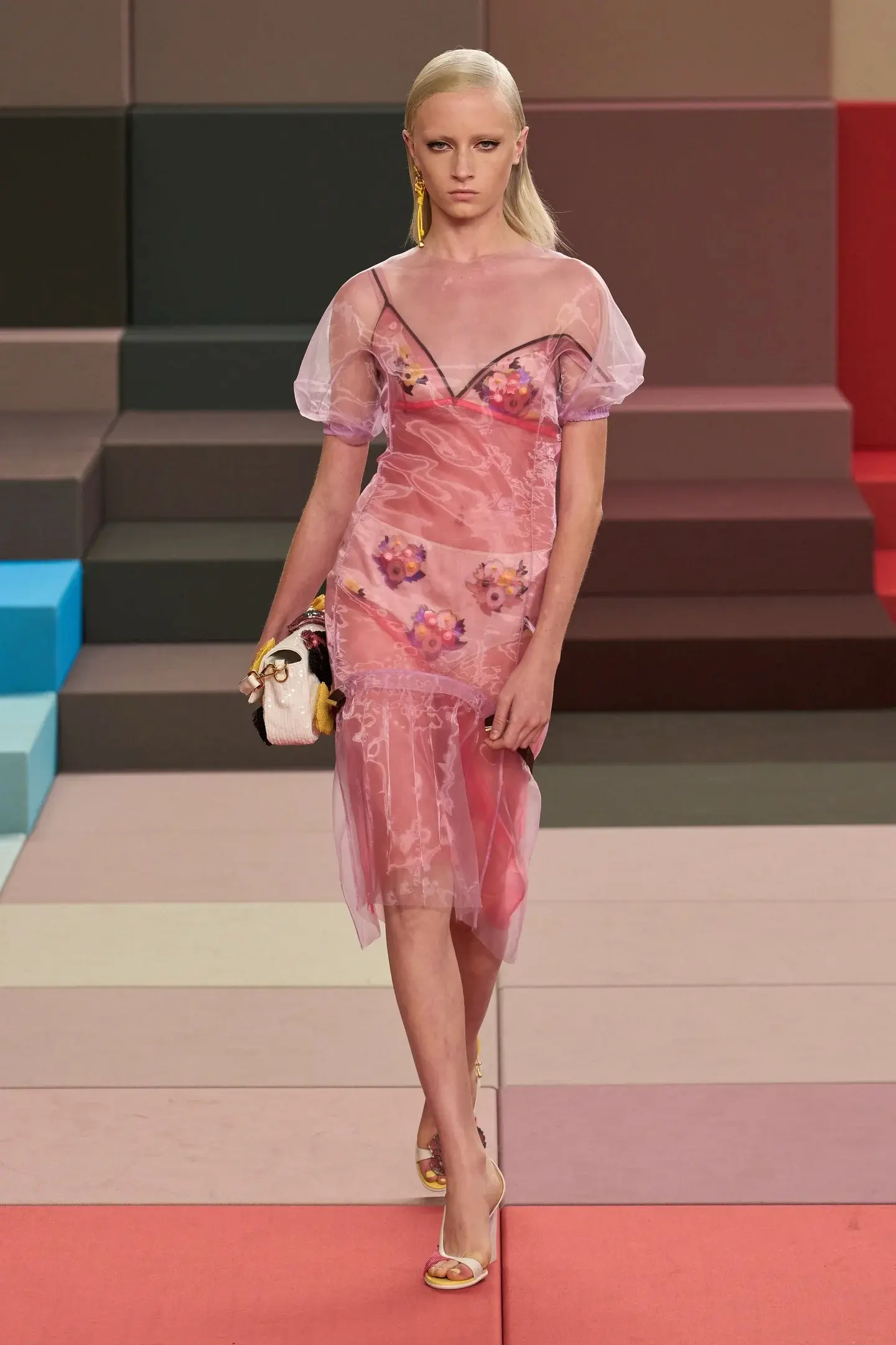 Modelo desfilando com vestido transparente de camadas em tons de rosa e flores, representando a tendência de transparência na moda SS26.