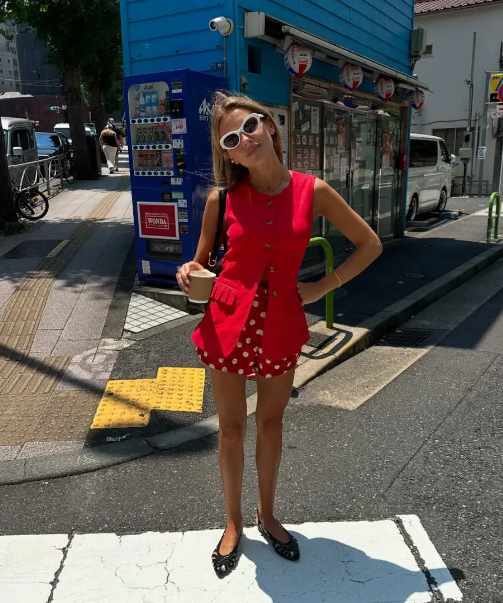 Mulher usando um colete vermelho com botões e shorts de poá, segurando um copo em uma rua de cidade ensolarada.