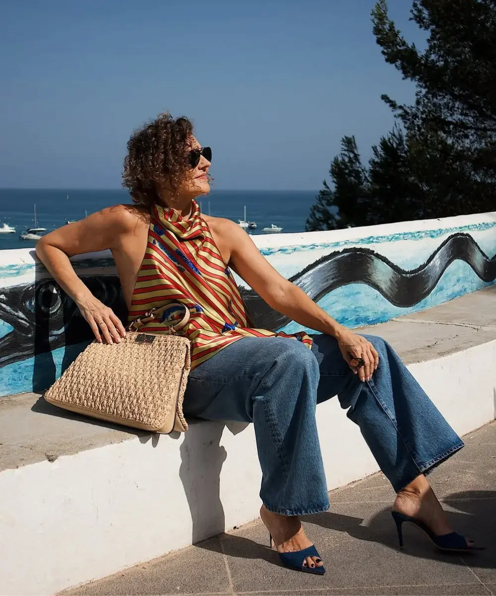 Mulher sentada ao sol, vestindo blusa acetinada listrada e calça jeans escura, segurando bolsa de palha.