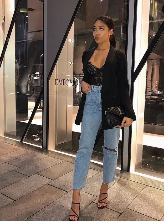 Mulher usando body de renda preto, blazer e jeans destroyed em look noturno elegante.