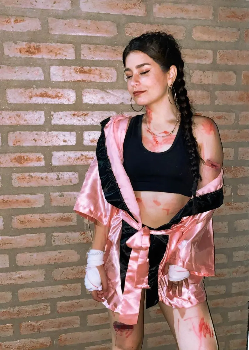 Mulher vestida com fantasia de boxeadora rosa e preta, com trança lateral e maquiagem simulando machucados.